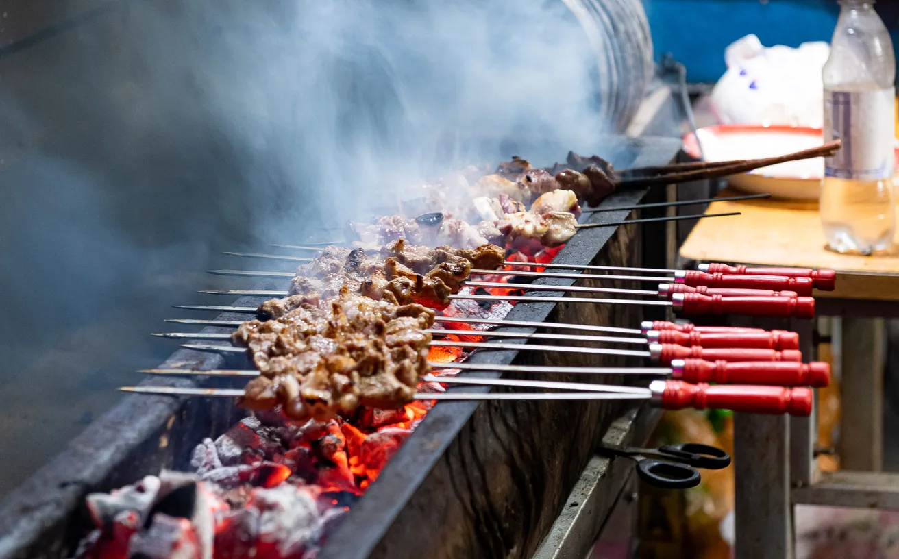Lamb kebabs on metal skewers over glowing charcoal in a long mangal grill, smoke rising, in the old city of Kashi